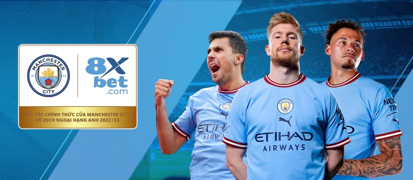 The image freezes the instant when 8Xbet and Manchester City football players come together, gathered in front of a banner that proudly announces their collaboration. The sportsmen are wearing their Manchester City jerseys and are placed on a football field. The banner prominently displays the logos of both 8Xbet and Manchester City